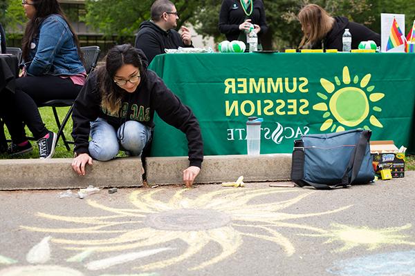 A photo of a student drawing on the pavement with chalk, in front of a table with a green banner reading 'Summer Session'